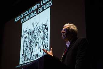 Photograph of someone speaking in front of a projected image onstage in the auditorium at Tate