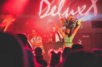 A person on stage in front of a crowd with musicians wearing a large colourful feather headpiece